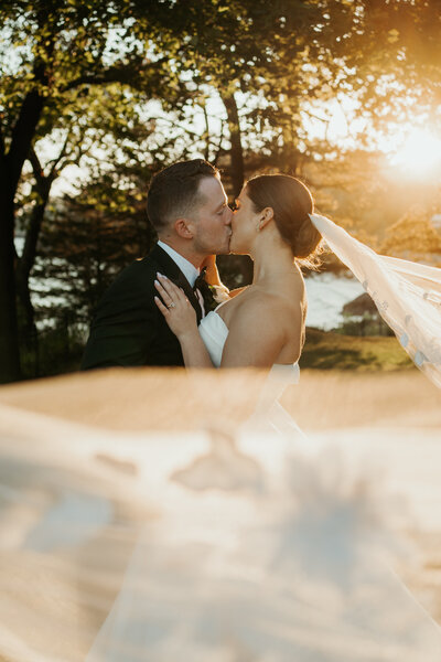 Bride & Groom share a romantic kiss as the bride's long veil blows in the wind and the golden sunset hits the veil just right to create a golden glow on the couple