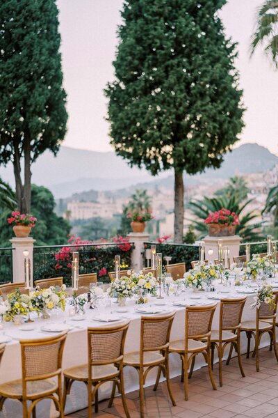 Long outdoor wedding dinner table with floral centerpieces and candlelight, set against a scenic mountain and cypress backdrop