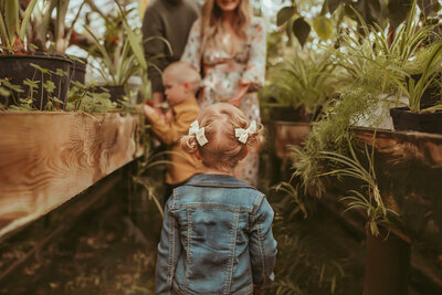 Little girl in blue jean jacket and blonde curly pigtails with bows walk away toward brother and mom in greenhouse family session