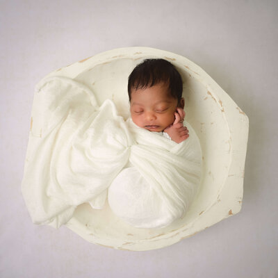 Newborn wrapped in white blanket and posed sleeping in a white bowl in an in-home newborn photo session in Bremerton, WA. Image by E. Benecki Photography.