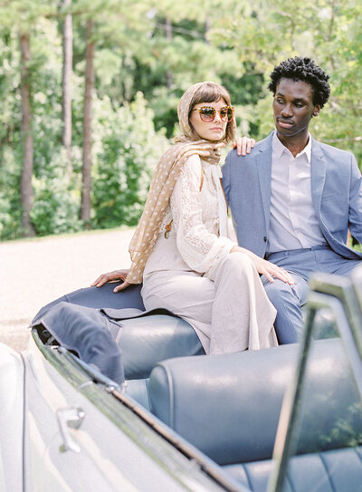 Engagement session with a 1940s vintage car. The car is a convertible and has pale blue interior. He is wearing the same shade blue suit with white dress shirt. She is in taupe colored wide legged dress pants with a cream colored lace shirt, oversized sunglasses and a tan and white polka dot scarf wrapped around her head and hanging down over her shoulder. She is looking ahead and he is looking at her. Photographed at River Oak with Low Country Valet car by wedding photographers in Charleston Amy Mulder Photography.