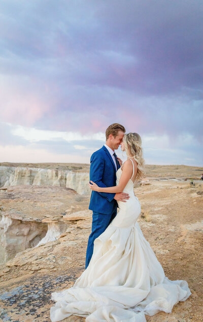 A bride and groom stand on the edge of the grand canyon as the have foreheads together and are smiling. There is a purple and pink sunset behind them.