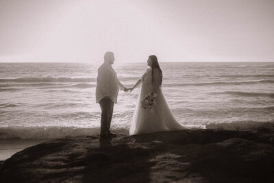 Bride and groom hold hands while facing each other in front of a sunset over the ocean. The bright sun creates sparkles on the water. The couple is standing on a large together. The bride holds her bouquet relaxed at her side and the groom has one hand in his pocket while he looks at his bride.