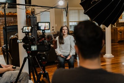 a woman with long black hair in a white blouse and blue jeans sits in front of cameras talking with video producer chris costello in the foreground.