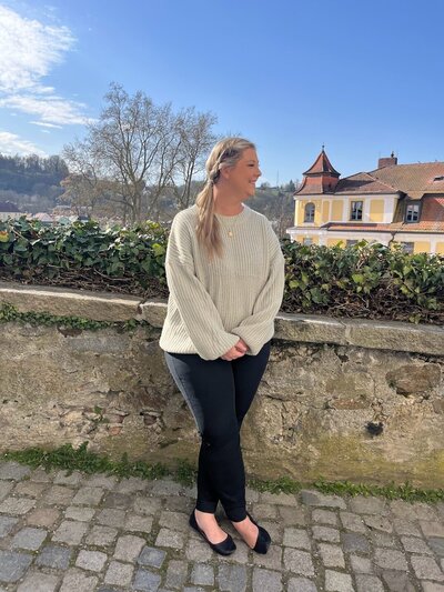 A woman with long hair stands smiling by a stone wall wrapped in green ivy. She's clad in a cream sweater, black pants, and black shoes. The picturesque backdrop features houses with red roofs under a clear blue sky, reminiscent of a perfect day from Epic Sands Travel.