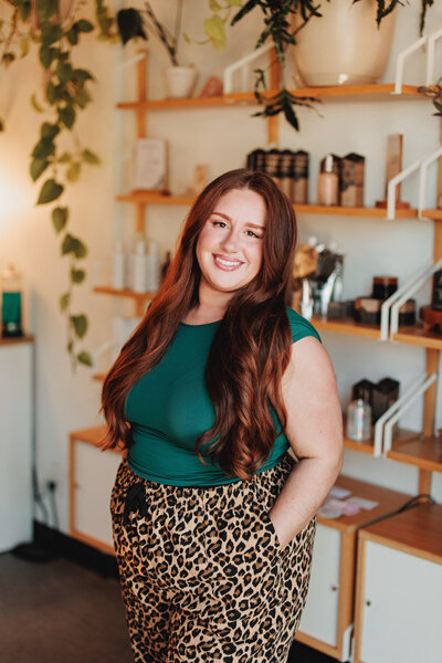 Morgan a hair stylist in Eugene stands in front of a wooden shelf with hair care products and smiles at the camera.