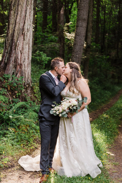 A blond groom wearing a dark gray suit and blush pink tie pulls his bride's chin in for a kiss as she holds her bouquet of white roses and greenery wearing a blush lace gown with sweetheart neckline standing in the lush forest of Southern Oregon near Ashland. | Erica Swantek Photography