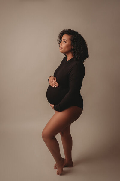 Atlanta, Georgia photographer photoshoot 3rd trimester pregnant woman posing in a Woodstock GA maternity portrait studio wearing a black long sleeve body suit  of her while she shows her baby bump. Shot on gray backdrop