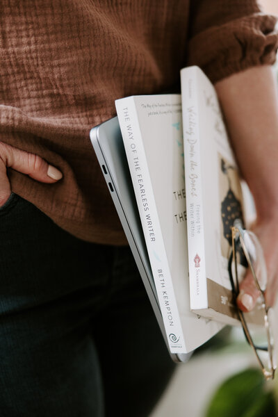 Freelance copywriter Erika Fitzgerald holding a MacBook, copywriting books, and reading glasses while wearing black pants and a brown blouse