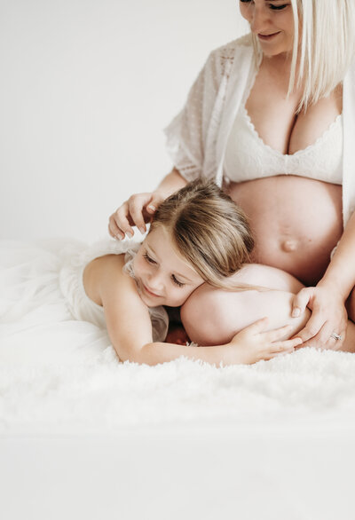 Young girl resting her head on her mother’s pregnant belly during an intimate in-home maternity session with a Kansas City maternity photographer.