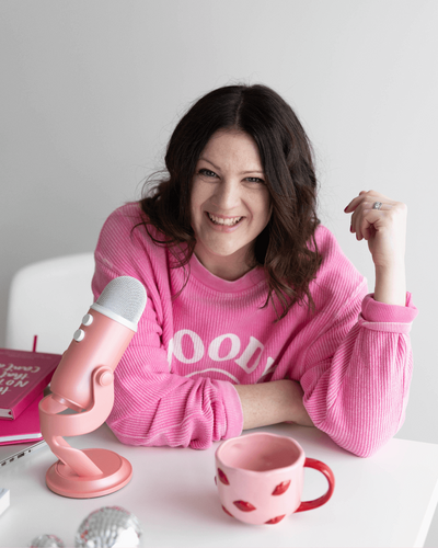 Emily, a brand and web designer for women, sits at a table in a pink sweatshirt, speaking into a microphone.