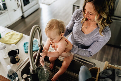 A mother holds her baby in a kitchen sink, gently guiding the baby’s hands under running water. The baby is curious, and the mother smiles as she supports the child.