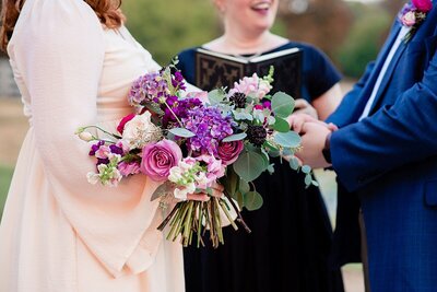 Bride in boho ivory dress and groom in bright blue suit say their vows while holding a large bouquet of pink and purple flowers