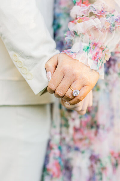 A stunning oval-cut engagement ring shines as a couple holds hands, framed by delicate floral details on the bride’s gown