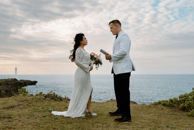 M facing J holding her bouquet as J reads vows to M standing on a cliffside views of the ocean