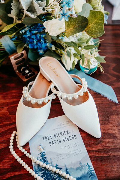 A detailed flat lay showcasing elegant white bridal shoes, wedding invitation, pearl jewelry, and a lush bouquet of greenery and blue flowers arranged artfully, capturing the details of a Lake Tahoe wedding