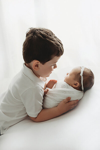 Big brother looks at his newborn baby sister in a white studio.