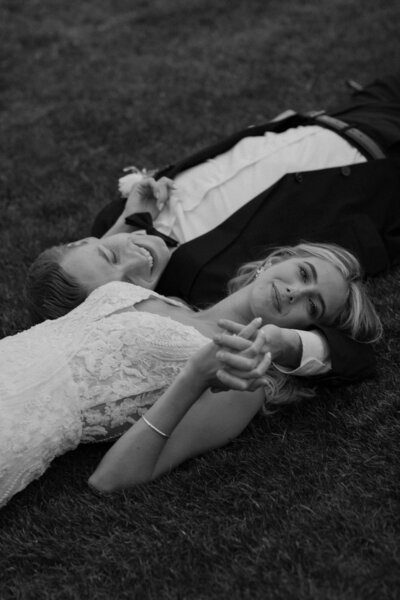 A newlywed couple laying opposite directions in the grass holding hands 