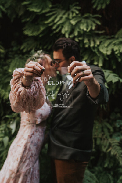 Bride and groom kissing while holding up we eloped sign in front of camera with a background of ferns by California elopement photographer Kasey Mantiply