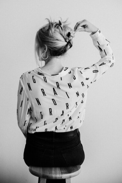 Black and white image of the back of Sarah Klongerbo sitting on a stool and pinning her hair up with pens