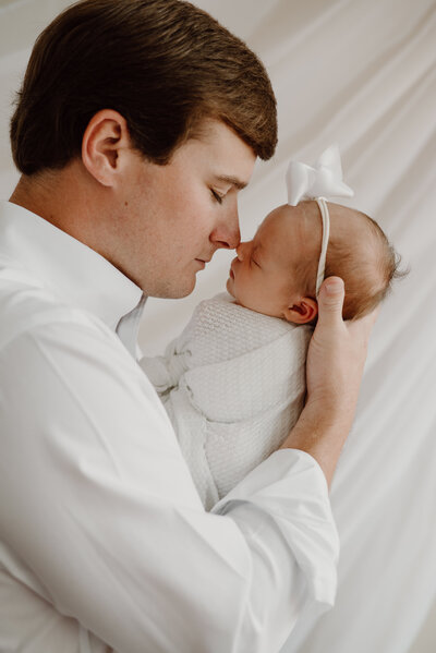 Newborn baby girl in white being held by dad in white and touching noses.