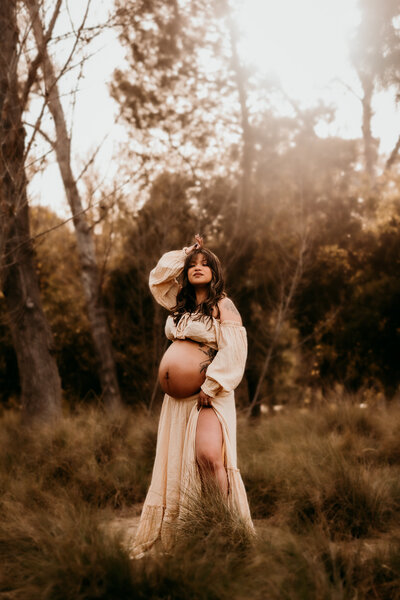 Pregnant woman in a cream dress holding her belly in a golden field during a maternity photoshoot in Corona, California, photographed by Brittani Luce

