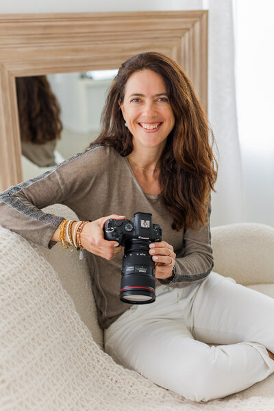 photographer woman wearing white jeans and beige top sitting on a sofa and holding a camera