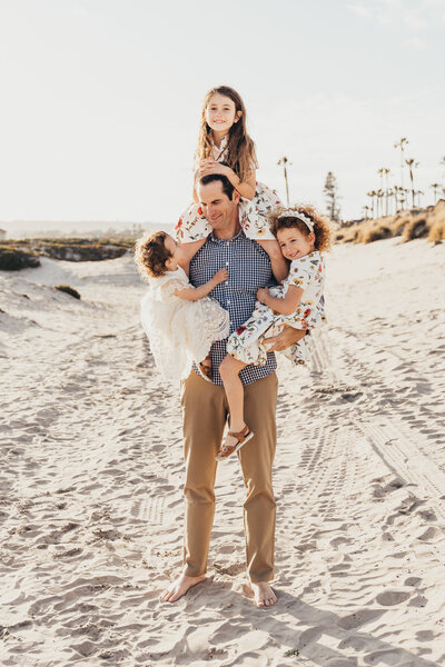 a dad holds his three daughters at one time while walking on the beach in Coronado San Diego