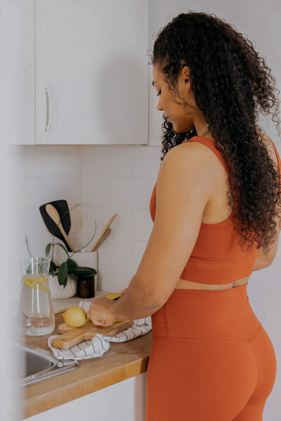 A woman with curly hair, in an orange sports outfit, stands at a kitchen counter slicing a lemon. A glass pitcher of lemon water hints at healthy habits—perfect for those seeking anxiety and depression counseling in Southern California.