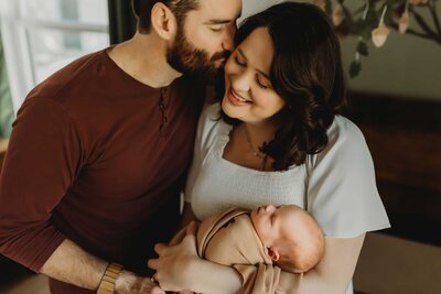 A tender moment of love and family during an intimate newborn home session, where a husband lovingly kisses his wife on the cheek. The mother is holding her new baby in her arms and the baby is sleeping while wrapped in a tan colored blanket.