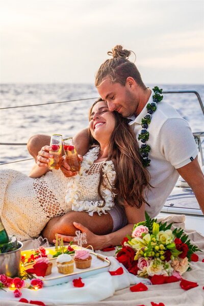 Engaged couple relaxing and embracing on a sailboat in Hawaii, clinking drinks and smiling with flowers and the ocean in the background during sunset.