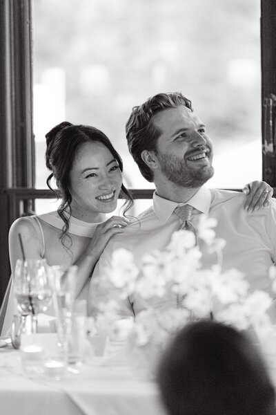 Candid black-and-white photo of bride and groom smiling during wedding toasts at their Colorado reception