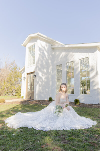 A bride sits on the grass in front of a large white wedding venue. She's posed and looks slightly editorial.