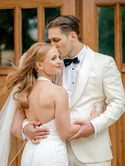 Groom in a white suit kisses bride, in a lace halter-neck gown, on the temple as he holds her she she smiles.