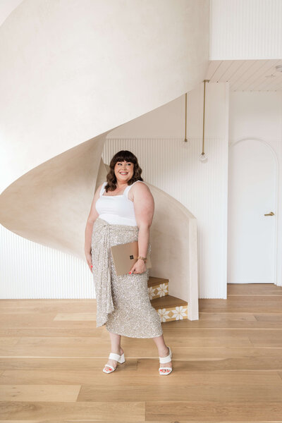Leith standing at the foot of a concrete staircase with laptop, wearing sequin skirt and white cami top