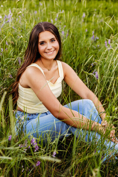 high school girl sitting as she hugs her knees while sitting in a field for her senior photos