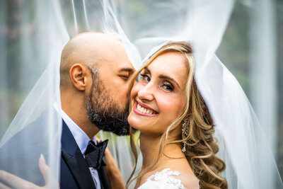 A bride wearing a white veil smiles while kissed on the cheek by her groom