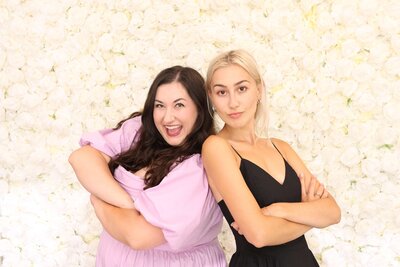 Clarae and Taressa, wedding planners in Oregon, stand in front of a wall of white roses with their backs against each other. 