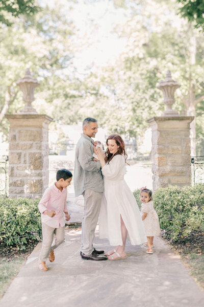 A family of five sits in a beautiful Kansas City garden. While mom holds her newborn, big sis looks on, while big brothers sits on his dad's lap who is lovingly smiling at him. 
