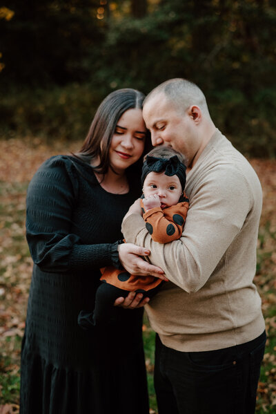 Mom in black dress and dad in tan shirt and black pants snuggling while dad is holding baby girl in orange and black dressin Annapolis Maryland.