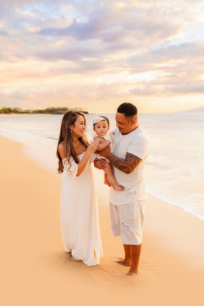 family of three with young toddler poses on the beach while looking at little girl