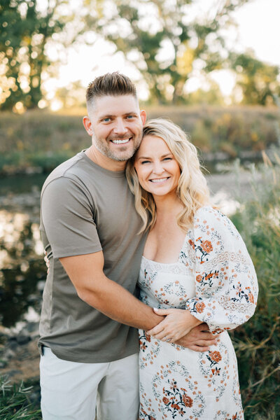 husband and wife hugging by the river and smiling at the photographer