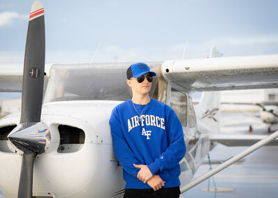 High school senior wearing a blue "Air Force AF" sweatshirt and matching cap, standing confidently in front of a small airplane with sunglasses on, under a clear sky.