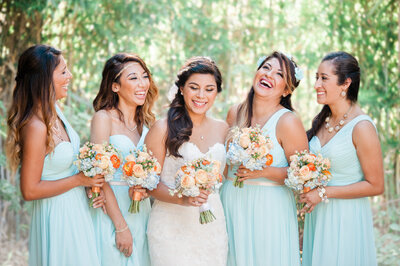 bride and her bridesmaids stand side by side holding bouquets laughing while getting ready for the wedding