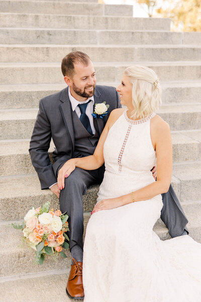 Bride and groom sitting next to each other on the Topeka Kansas Capitol steps with a fall colored bouquet sitting next to them by Kansas City Wedding Photographer Sarah Riner Photography.