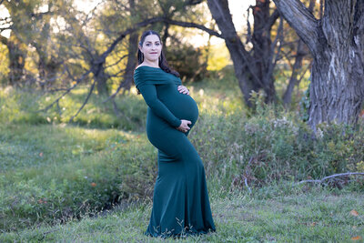 Maternity photo in Dallas Fort Worth of a woman in a green dress in a grassy field by a tree showing off her bump