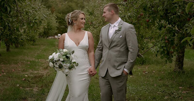 Couple standing side- by- side. holding hands in an apple orchard. Bride is wearing a low cut white dress with lace accents. She has a green and whire bouquet in her right hand and has a cathedral length veil. Groom is wearing a grey suit with a white tie and is standing with his left hand in his pocket