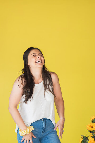 Bee is smiling with her eyes closed, mouth wide open as if she's laughing, she stands in front of a yellow backdrop wearing a casual outfit of jeans and a tank top