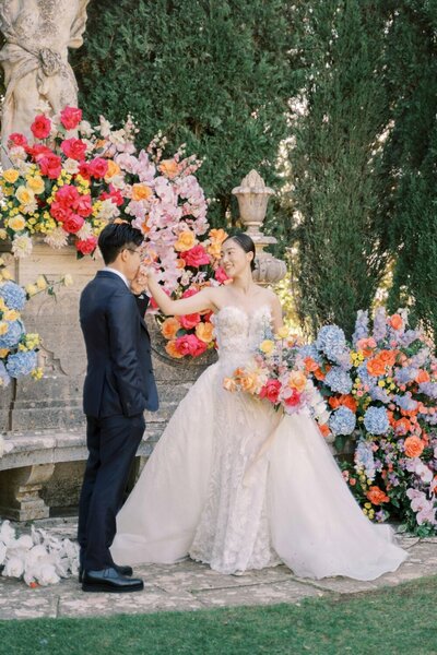 A bride and groom embrace joyfully in a french chateau on a sunny day, captured beautifully by Andreas K. Georgiou, international wedding photographer. The bride wears a white dress with lace sleeves and a floral veil, while the groom is in a suit holding a small bouquet. Sunlight creates a warm glow around them.