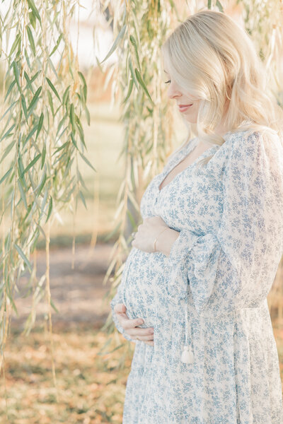 Pregnant mother holds her baby bump while looking down in a blue floral dress in front of willow trees in Indianapolis